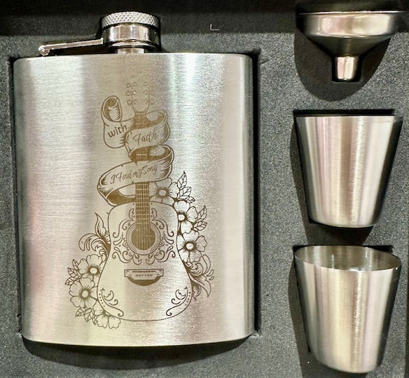 Stainless steel flask set with engraved design, two shot glasses, and a funnel on a dark surface.